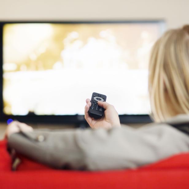 Blonde on red couch points remote control at TV