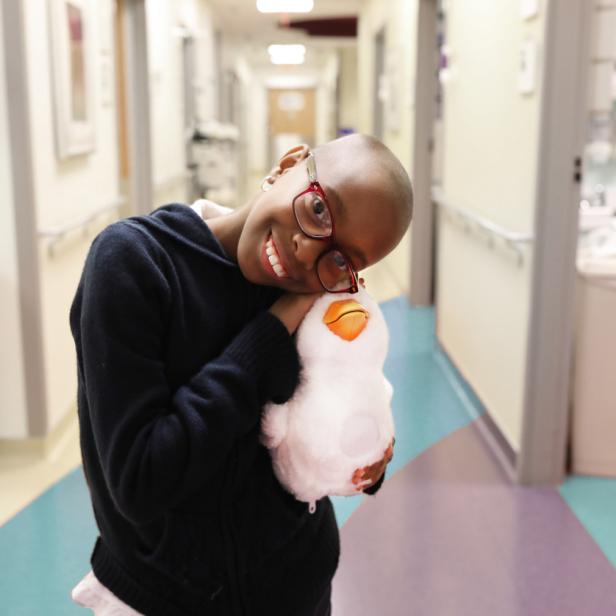 Ein lächelndes Mädchen mit Brille umarmt ein Stofftier in einem Krankenhausflur.