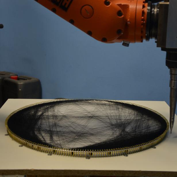 Ein Roboterarm bearbeitet ein rundes Objekt, das mit schwarzen Fäden bespannt ist.