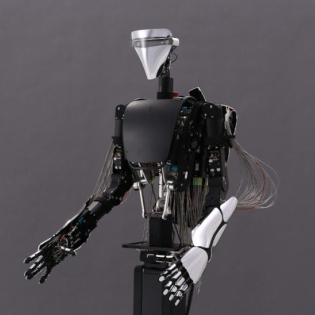 Ein humanoider Roboter mit freiliegenden Kabeln und mechanischen Details vor grauem Hintergrund.