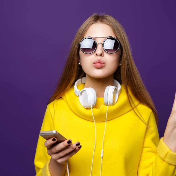 Portrait young girl teenager with earphones and phone, in a yellow sweater, isolate on a violet background.