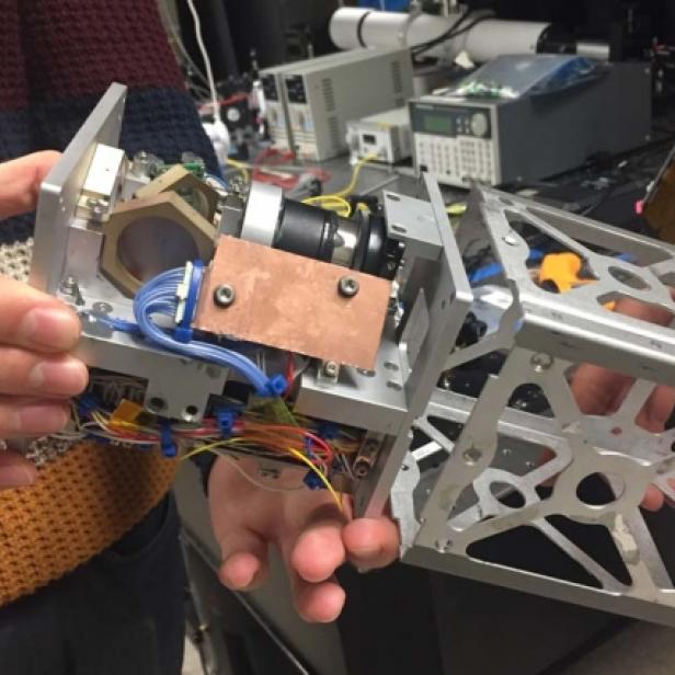 Zwei Personen halten ein komplexes, silberfarbenes Gerät mit Kabeln und optischen Elementen.