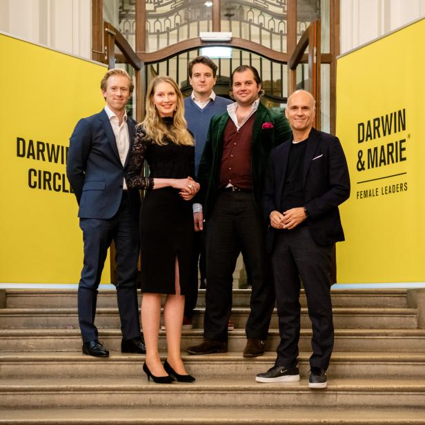 Eine Gruppe von fünf Personen posiert auf einer Treppe vor Bannern von „Darwin & Marie“.