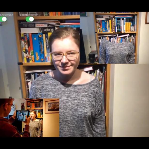 Eine Frau mit Brille steht vor einem Bücherregal, während sie von einem Tablet fotografiert wird.