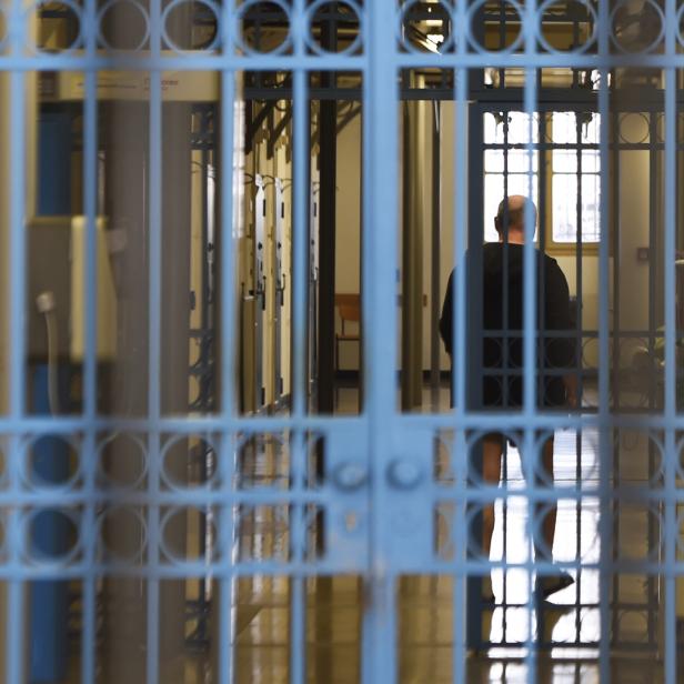 Eine Person steht hinter blauen Gitterstäben in einem Flur mit Zellentüren und Fenstern.