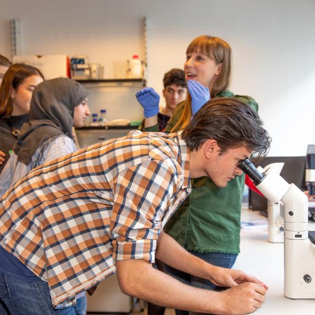 Ein junger Mann blickt durch ein Mikroskop, umgeben von einer Gruppe weiterer Personen.