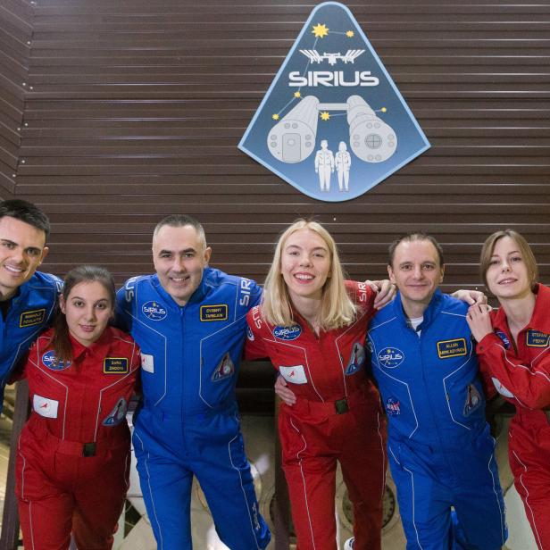 Eine Gruppe von sechs Personen in blauen und roten Raumanzügen posiert vor einem SIRIUS-Logo.