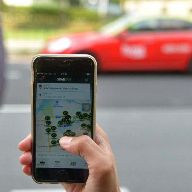 Eine Person benutzt die GrabTaxi-App auf einem Smartphone, um eine Fahrt zu buchen. Im Hintergrund ist ein rotes Auto zu sehen.