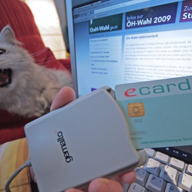Eine e-card wird vor einem Laptop mit einer Webseite zur ÖH-Wahl und einer Katze im Hintergrund gehalten.