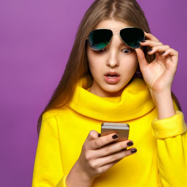 Portrait surprised young girl from message on smartphone in brightly yellow sweater, isolate on a violet background