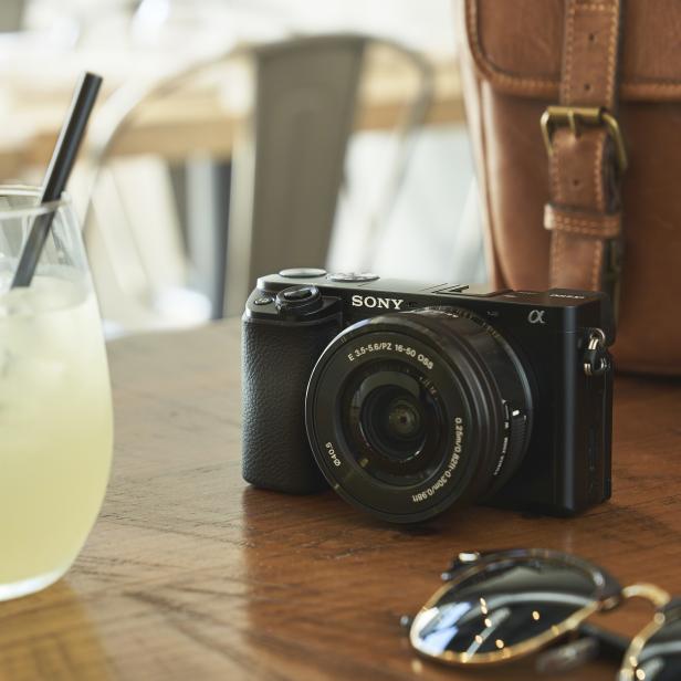 Ein Cocktail mit Limette steht neben einer Sony-Kamera und einer braunen Ledertasche auf einem Holztisch.