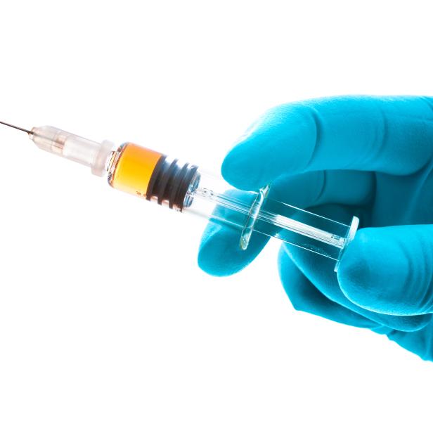 Eine Hand in einem blauen Handschuh hält eine Spritze mit einer gelblichen Flüssigkeit.