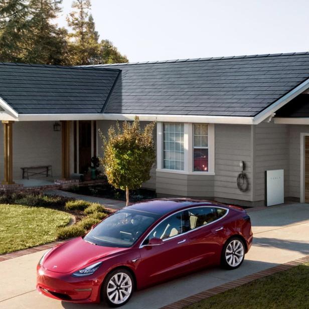Ein rotes Tesla Model 3 steht vor einem Haus mit Solardach und Tesla Powerwall.