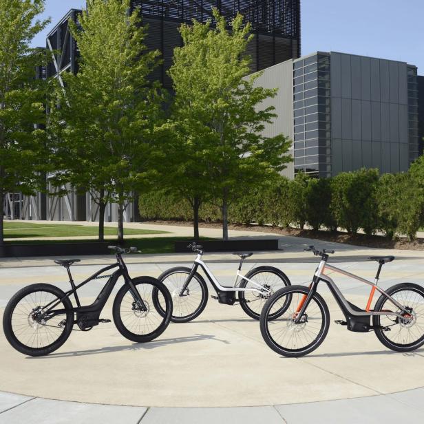 Drei E-Bikes in Schwarz, Weiß und Orange stehen auf einem Platz vor modernen Gebäuden.