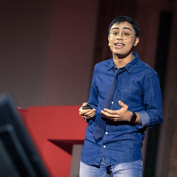 Ein junger Mann hält einen Vortrag auf einer Bühne mit rotem Hintergrund.