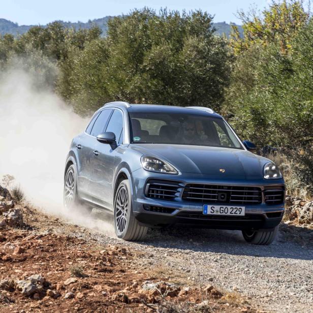 Ein blauer Porsche Cayenne fährt auf einer unbefestigten Straße und wirbelt Staub auf.