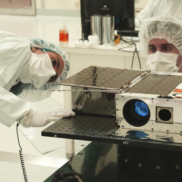 Zwei Personen in Reinraumkleidung inspizieren einen Satelliten mit Solarzellen.