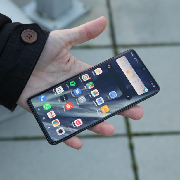 Eine Hand hält ein Smartphone, auf dessen Bildschirm Apps und Wetterinformationen für Wien angezeigt werden.