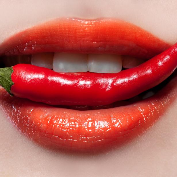 Nahaufnahme von einem Mund mit rotem Lippenstift, der eine rote Chili zwischen den Zähnen hält.