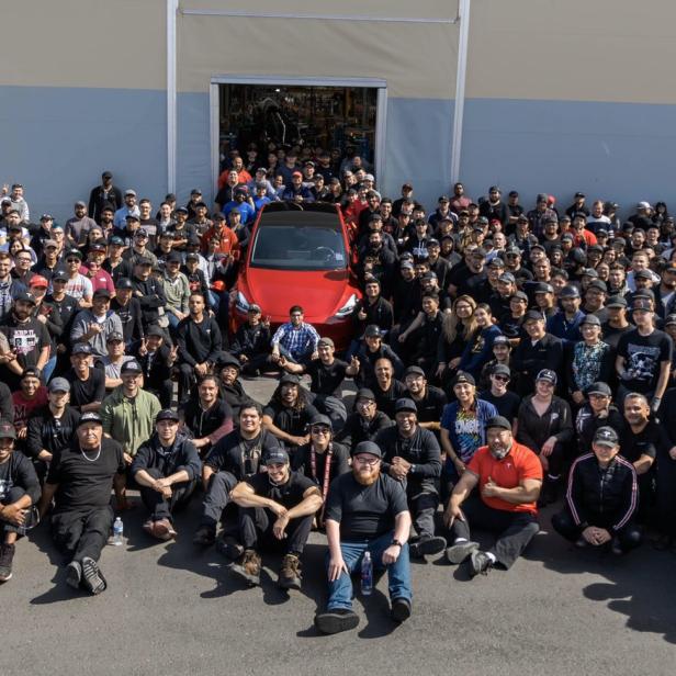 Eine große Gruppe von Tesla-Mitarbeitern posiert vor einem roten Tesla Model Y vor einem Lagerhaus.