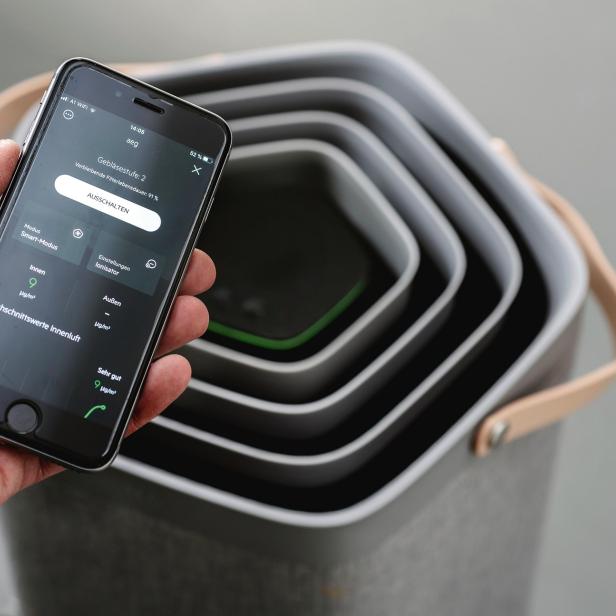 Eine Hand hält ein Smartphone vor einen Luftreiniger, auf dem die zugehörige App geöffnet ist.
