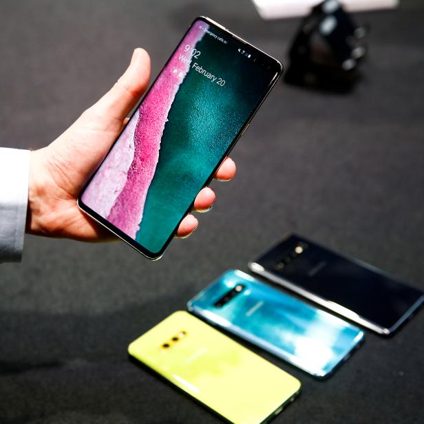 Eine Hand hält ein Smartphone mit farbigem Display; weitere Smartphones liegen auf einem Tisch.