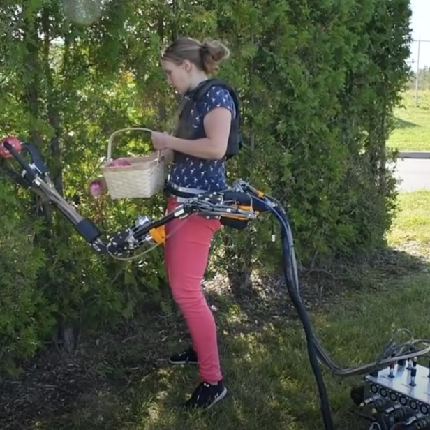 Eine Frau pflückt mit einem Roboterarm Äpfel in einen Korb, während ein Mann die Steuerung bedient.