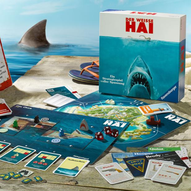 Das Brettspiel „Der Weiße Hai“ mit Spielbrett, Karten, Würfeln und einer Haifischflosse im Hintergrund.