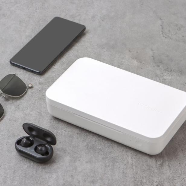 Ein Smartphone, eine UV-Sterilisationsbox, Ohrhörer und eine Sonnenbrille liegen auf einer grauen Oberfläche.