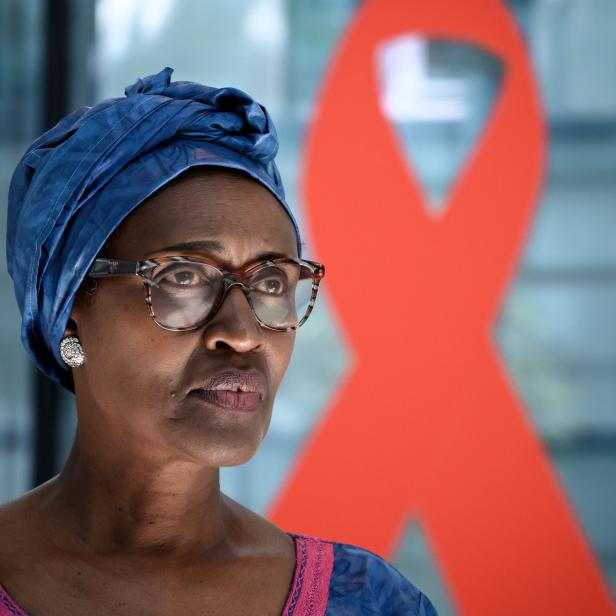 Eine Frau mit blauem Kopftuch und Brille vor einer roten Schleife, dem Symbol für den Kampf gegen AIDS.