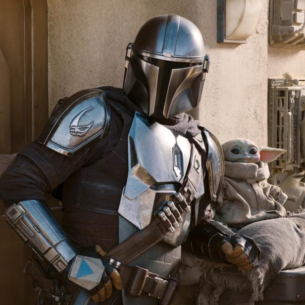 Der Mandalorianer mit Baby Yoda im Arm, beide vor einer Wand lehnend.