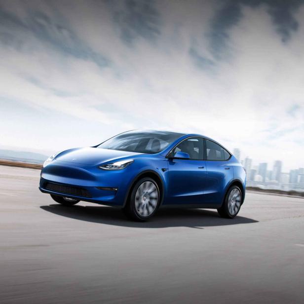 Ein blaues Tesla Model Y fährt auf einer Straße mit einer Stadt im Hintergrund.