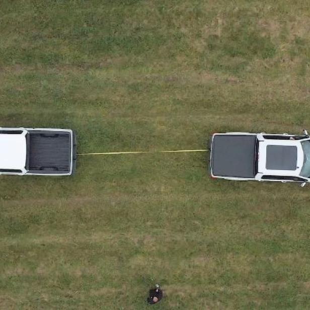 Zwei weiße Pick-up-Trucks stehen auf einer Wiese und sind mit einem gelben Seil verbunden.