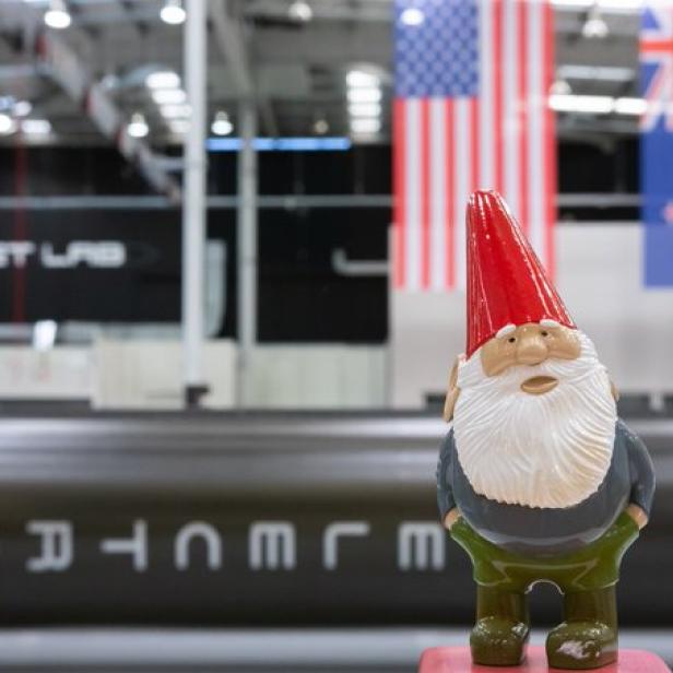 Ein Gartenzwerg steht vor einer Rakete im Rocket Lab, mit Flaggen der USA und Neuseelands im Hintergrund.