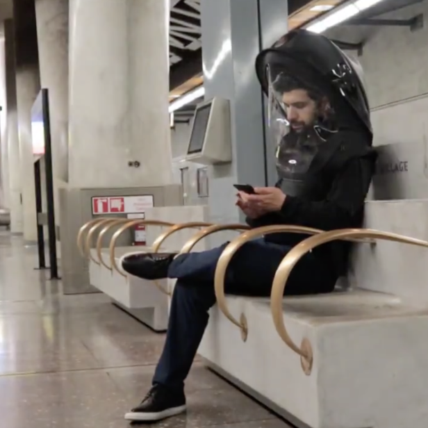 Ein Mann mit einem großen, schützenden Helm sitzt auf einer Bank und benutzt sein Handy.
