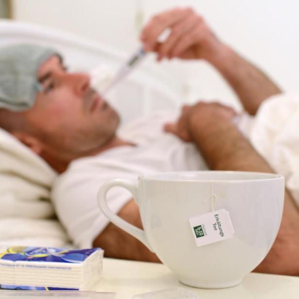 Ein kranker Mann liegt mit einem Thermometer im Bett, neben ihm eine Tasse Tee und Taschentücher.
