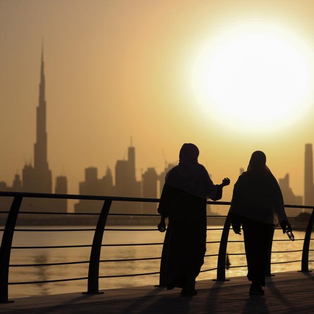 Zwei Frauen in traditioneller Kleidung spazieren vor der Skyline von Dubai in der Abenddämmerung.