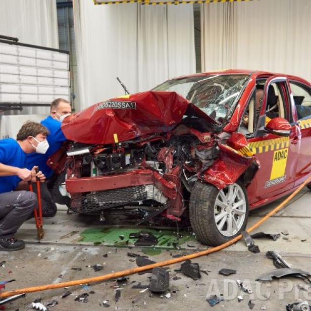 Ein rotes Auto nach einem Crashtest, untersucht von zwei Technikern mit Masken.