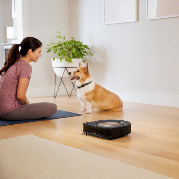 Eine Frau, ein Corgi und ein Staubsaugerroboter in einem hellen Raum.