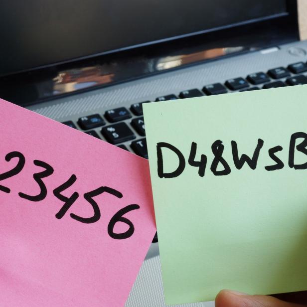 Eine Hand hält zwei Zettel mit den Passwörtern „123456“ und „D48WsB+2“ vor einem Laptop.