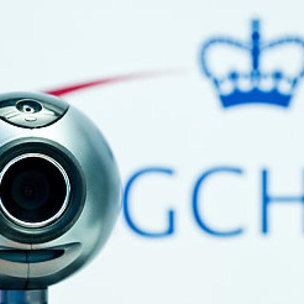 Eine silberne Webcam vor dem Logo des GCHQ, einer britischen Geheimdienstorganisation.