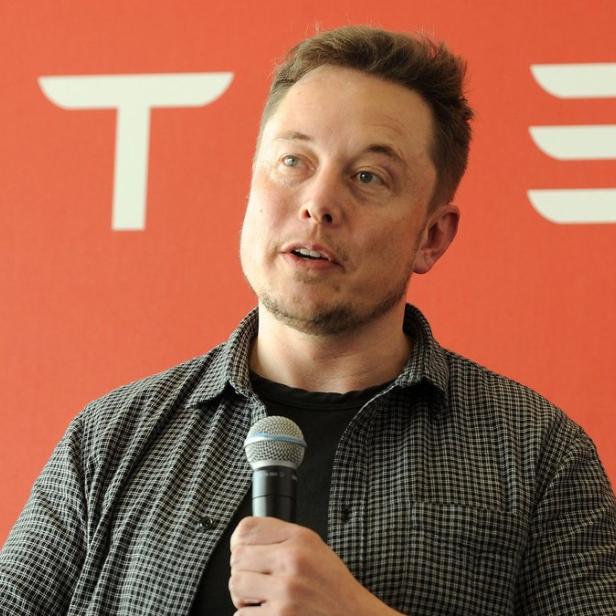 Elon Musk spricht vor einem roten Hintergrund mit dem Tesla-Logo.