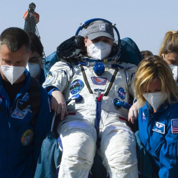 Ein Astronaut in seinem Sitz wird von mehreren Personen mit Masken umgeben.