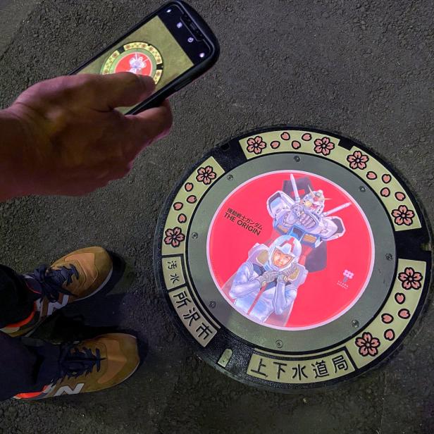 A passerby using a smartphone takes a photo of an illuminated manhole cover with designs of popular animation character Gundam, on the street in Tokorozawa, near Tokyo