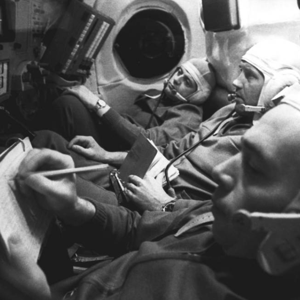 Drei Astronauten in Raumanzügen sitzen in einem Raumschiff, einer schreibt in ein Notizbuch.