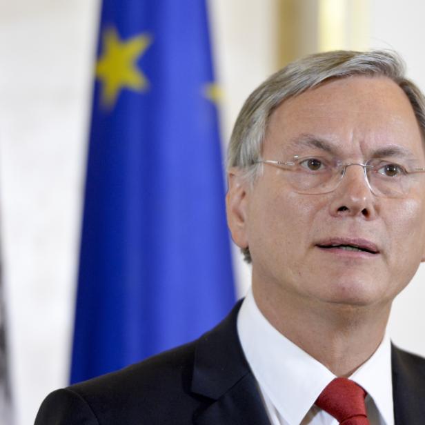 Ein Mann mit Brille und Anzug steht vor der österreichischen und der EU-Flagge.