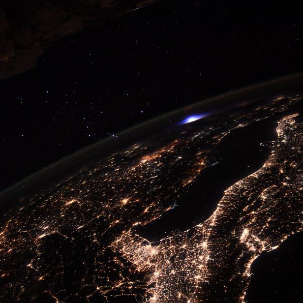 Blick aus dem Weltraum auf die Erde bei Nacht mit Lichtern und einem blauen Blitz.