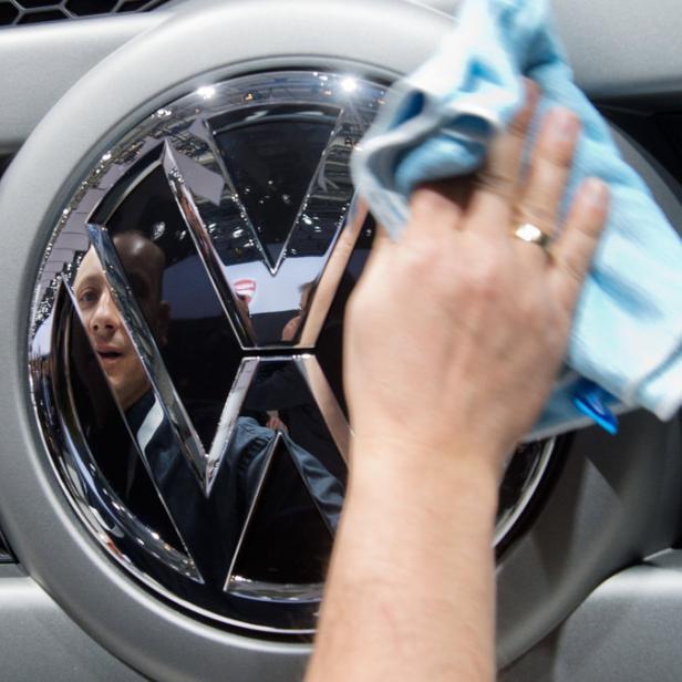 Eine Hand poliert mit einem blauen Tuch das VW-Logo an einem grauen Auto.