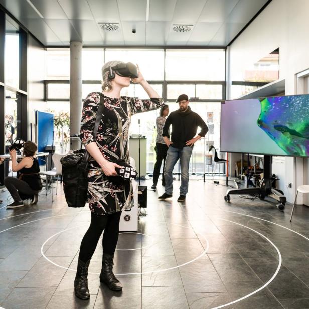 Eine Frau mit VR-Brille steht in einem Raum mit anderen Personen und einem großen Bildschirm.