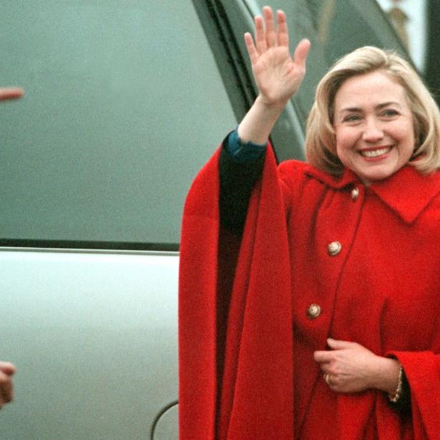 Hillary Clinton in einem roten Mantel winkt lächelnd.